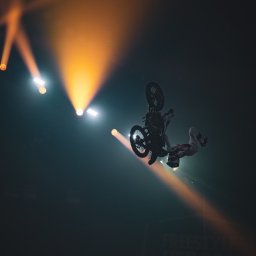 FQ Fotografia - Motocyklista wykonuje trik w powietrzu podczas pokazu freestyle motocross, oświetlony reflektorami na ciemnym tle.