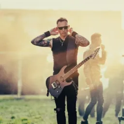 Gitarzysta z tatuażami, w czarnych okularach i ubraniach, trzymający gitarę elektryczną na tle sceny podczas koncertu na świeżym powietrzu, z delikatną poświatą.