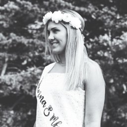 FQ Fotografia - Czarno-białe zdjęcie uśmiechniętej panny młodej w białej sukience z koronką, z wiankiem z białych kwiatów na głowie i szarfą z napisem 'Przyszła Młoda', pozującej na tle drzew.