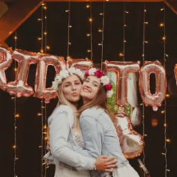 Dwie uśmiechnięte kobiety w wiankach z kwiatów, obejmujące się na tle balonów w kolorze różowego złota z napisem 'Bride to be' i delikatnych lampek.