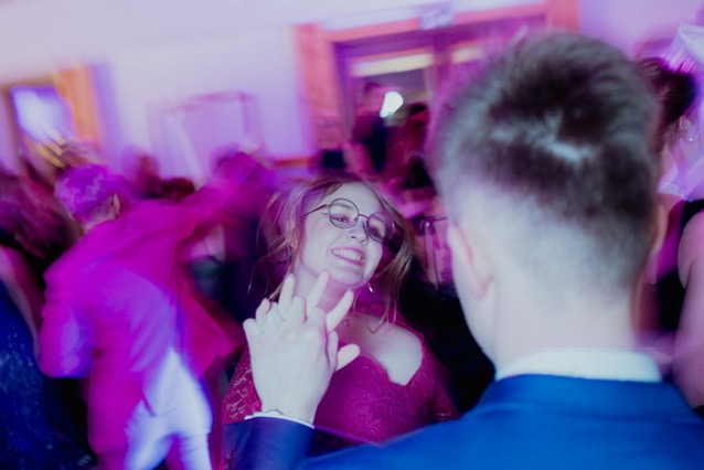 Uśmiechnięta kobieta w okularach, w czerwonej sukience, tańcząca na imprezie z niebieskim oświetleniem, widok zza ramienia mężczyzny w niebieskim garniturze.