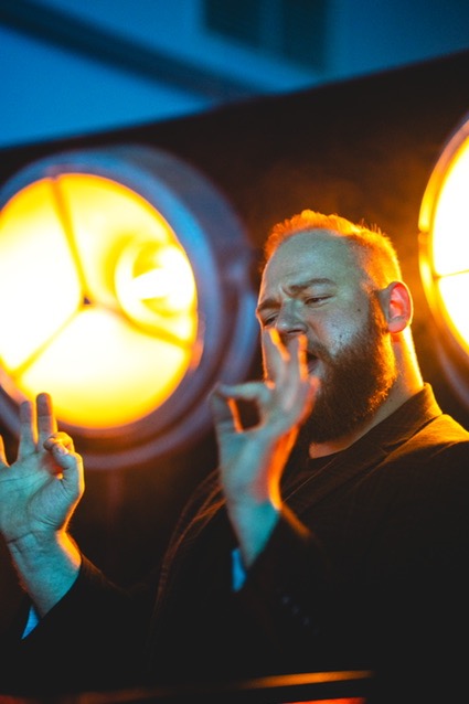 Kamerzysta z brodą, w czarnym garniturze, wykonuje gest dłońmi na tle okrągłych lamp o ciepłym świetle podczas filmowania wydarzenia.