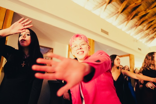 Rozmazane ujęcie z imprezy z młodą osobą w różowym garniturze i okularach, wyciągającą rękę w stronę kamery, w tle tańczący tłum.