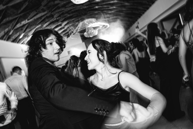 Czarno-białe zdjęcie pary tańczącej na parkiecie, widoczny tłum ludzi w tle i dekoracyjne oświetlenie sufitowe.