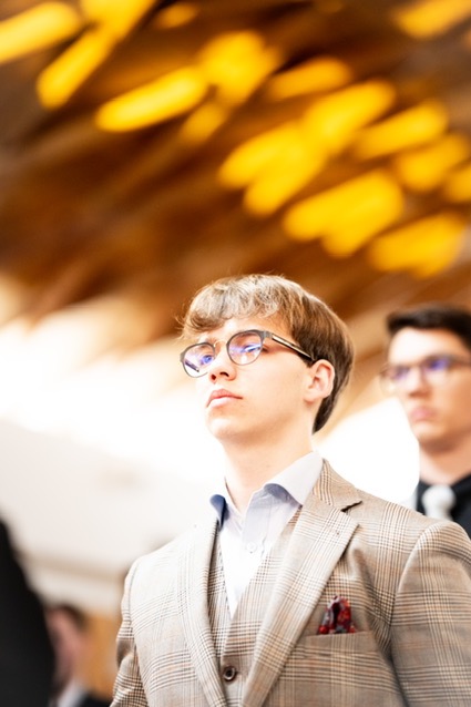 Portret młodego mężczyzny w okularach i kraciastej, beżowej marynarce z poszetką, spoglądającego w górę pod dekoracyjne oświetlenie.