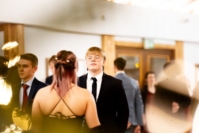 Mężczyzna w czarnym garniturze i okularach, otoczony innymi osobami na uroczystości, widziany zza pleców kobiety w czarnej sukni z cienkimi ramiączkami, z rozproszonym światłem w pierwszym planie.