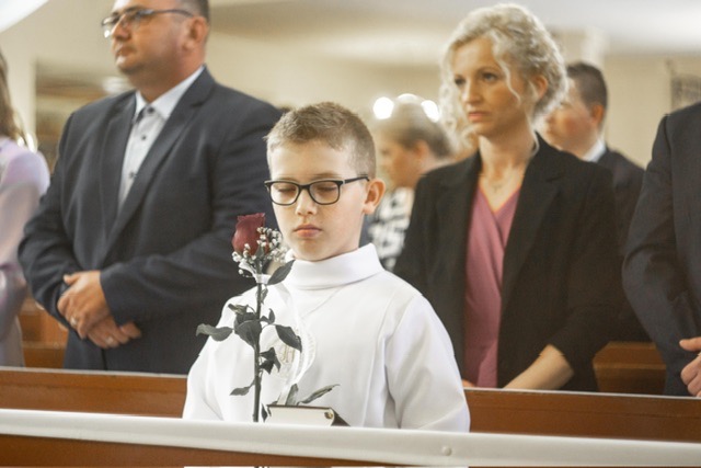 Chłopiec w stroju komunijnym, trzymający czerwoną różę w kościele, z zamyślonym wyrazem twarzy, w tle widoczni rodzice i inni uczestnicy uroczystości.