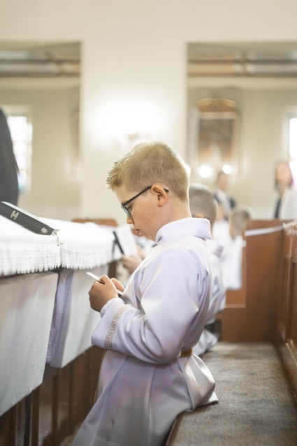 Chłopiec w albie i okularach klęczy w kościele, trzymając kartkę podczas uroczystości religijnej.
