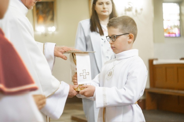 Chłopiec w okularach, ubrany w białą albę z haftowanym symbolem, trzyma złoty kielich z białą stułą z wyhaftowanym krzyżem podczas ceremonii religijnej w kościele, w tle kobieta w jasnym żakiecie.