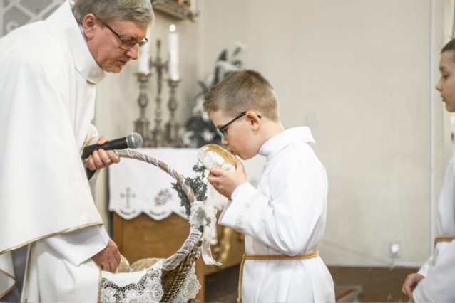 Chłopiec w albie z okularami całuje chleb podczas ceremonii religijnej, ksiądz trzyma mikrofon i koszyk z chlebem, widoczne dekoracje kwiatowe.