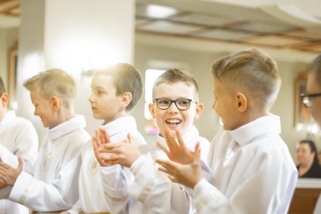 Chłopcy w białych albach klaszczą podczas uroczystości Pierwszej Komunii Świętej, jeden z chłopców uśmiecha się szeroko w okularach.