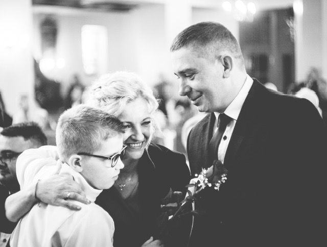 Czarno-białe zdjęcie z uroczystości rodzinnej: uśmiechnięta kobieta obejmuje chłopca w okularach, obok stoi mężczyzna w garniturze z kwiatem w ręku.