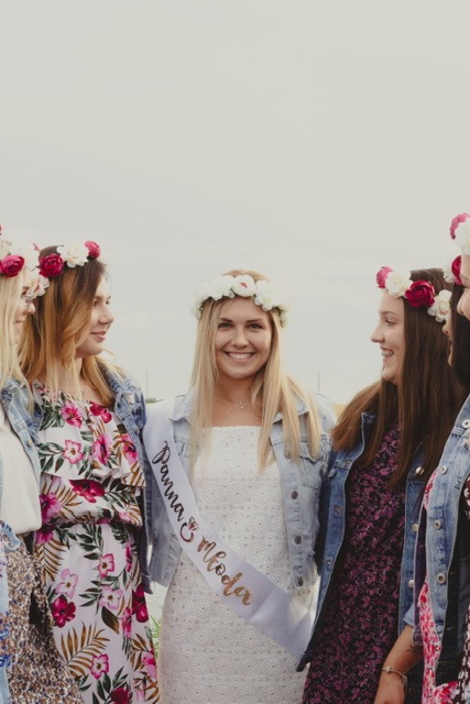 Panna młoda w białej sukience i wianku z białych kwiatów, otoczona przez druhny w denimowych kurtkach i wiankach z czerwonych i białych kwiatów, podczas wieczoru panieńskiego.