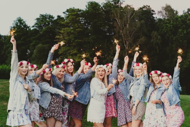 Grupa kobiet w kwiecistych wiankach i dżinsowych kurtkach świętuje, trzymając w górze odpalone zimne ognie na tle zieleni.