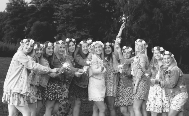 Czarno-białe zdjęcie grupy kobiet w wiankach na głowach, trzymających zapalone zimne ognie na tle zieleni.