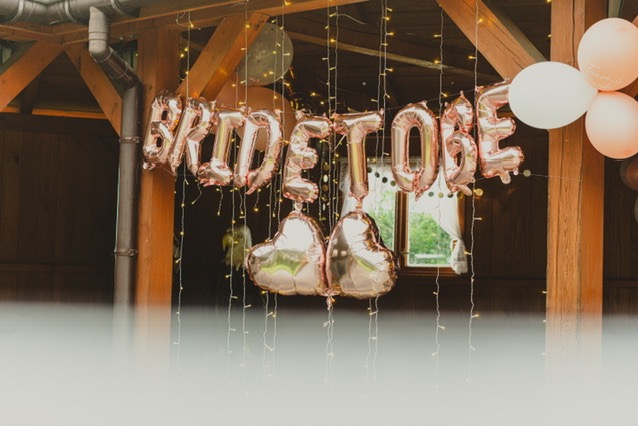 Dekoracja z różowych balonów foliowych 'Bride to be' i balonów w kształcie serc, zwisające lampki na tle drewnianej konstrukcji z oknem.