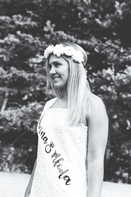 Czarno-białe zdjęcie uśmiechniętej panny młodej w białej sukience z koronką, z wiankiem z białych kwiatów na głowie i szarfą z napisem 'Przyszła Młoda', pozującej na tle drzew.