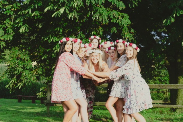 Grupa kobiet w kwiecistych sukienkach i wiankach na głowach, stojących w kręgu na trawie w parku, z drzewem w tle, ujęcie z delikatnym, naturalnym światłem.