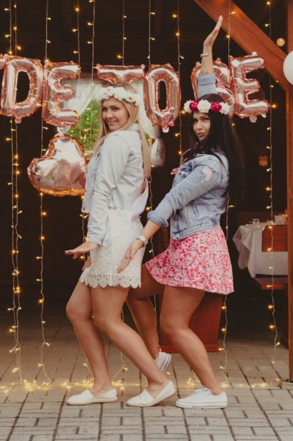 Dwie uśmiechnięte kobiety w wiankach i dżinsowych kurtkach pozują do zdjęcia na tle dekoracji z balonów Bride To Be i lampek w Toszku, prawdopodobnie podczas wieczoru panieńskiego.