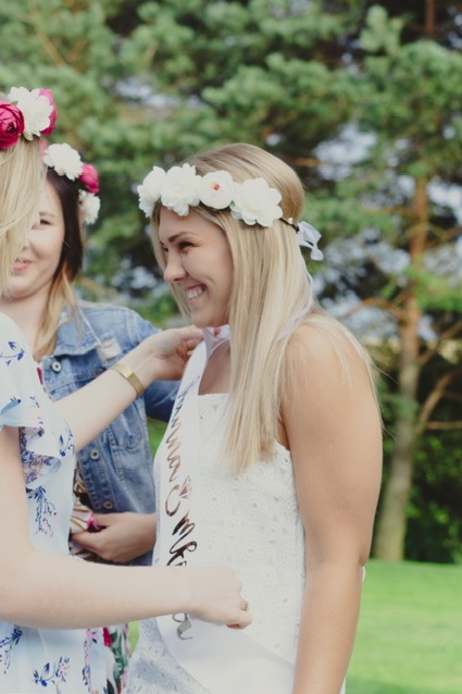 Uśmiechnięta blondynka z wiankiem z białych kwiatów na głowie, ubrana w białą sukienkę, podczas przygotowań do wieczoru panieńskiego; szarfa z napisem 'Bride to be' zakładana przez inną kobietę.