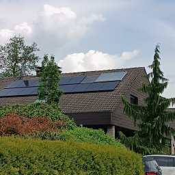 Alpha Pover - Brązowy dach domu z panelami słonecznymi, widok zza gęstego żywopłotu, z flagą w tle.