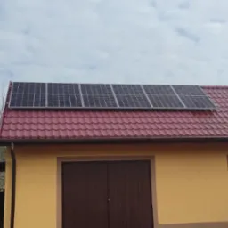 Panele słoneczne zamontowane na czerwonym dachu budynku z żółtą elewacją, widok z dołu, lekko pochmurne niebo.