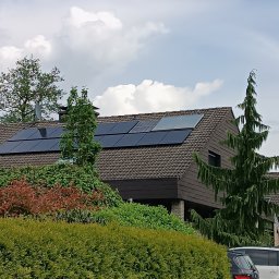 Alpha Pover - Panele słoneczne na brązowym dachu domu jednorodzinnego, widok z poziomu ulicy, zieleń w tle i flaga na maszcie.