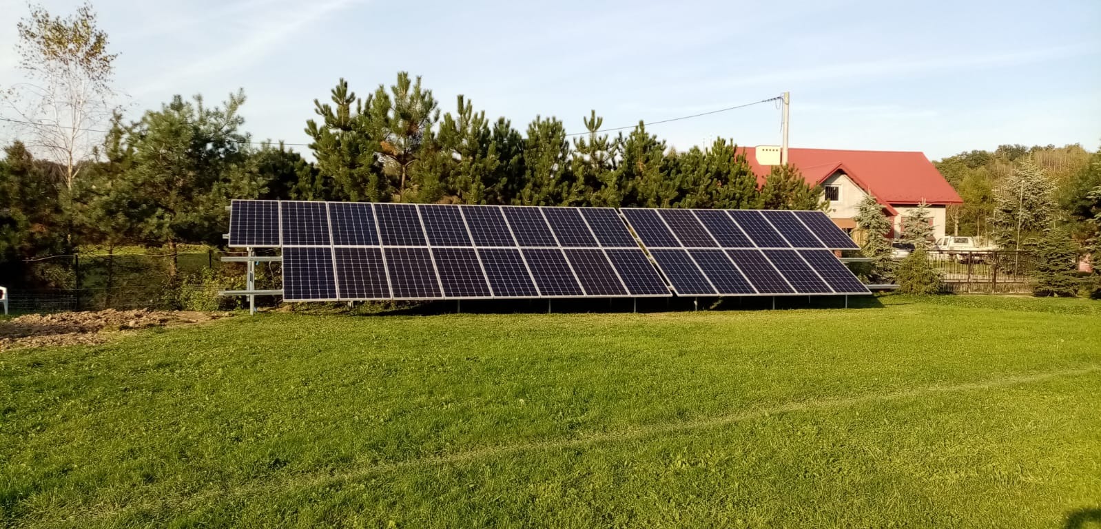 Instalacja fotowoltaiczna, panele na metalowej konstrukcji ustawione na trawniku przed domem jednorodzinnym z czerwonym dachem, widok z daleka.