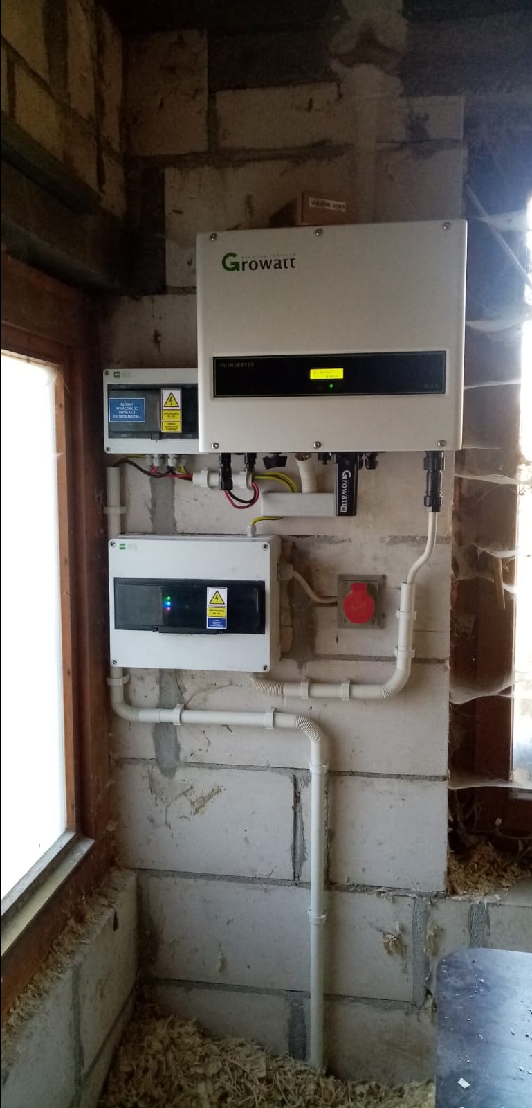Zamontowany inwerter fotowoltaiczny Growatt z rozdzielnicą i zabezpieczeniami na surowej ścianie z bloczków, widoczne przewody i rurki instalacyjne.