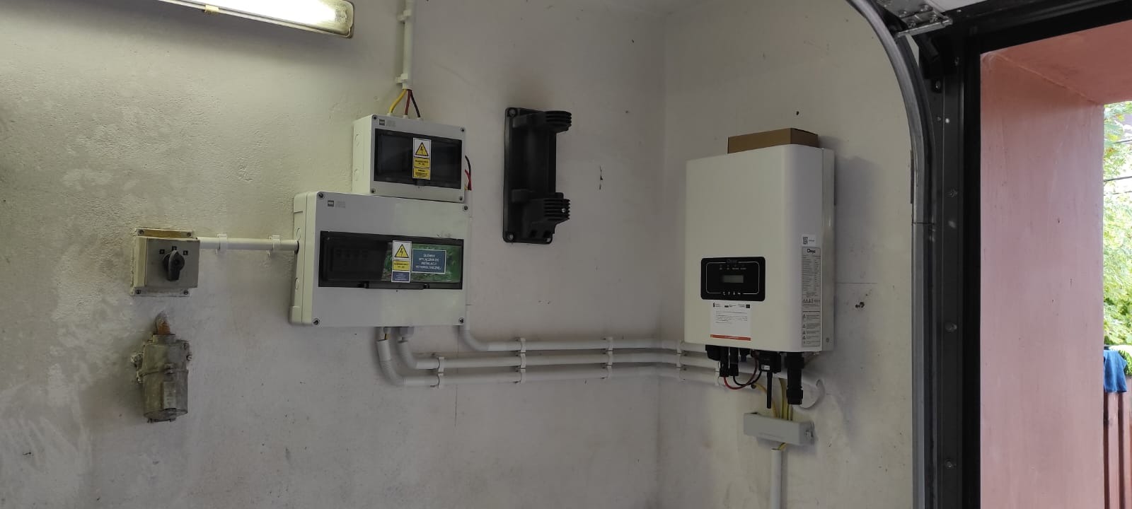 Instalacja elektryczna z falownikiem przy garażu: skrzynki bezpiecznikowe, rury PCV, biały falownik z wyświetlaczem, widoczne kable i przyłącza.