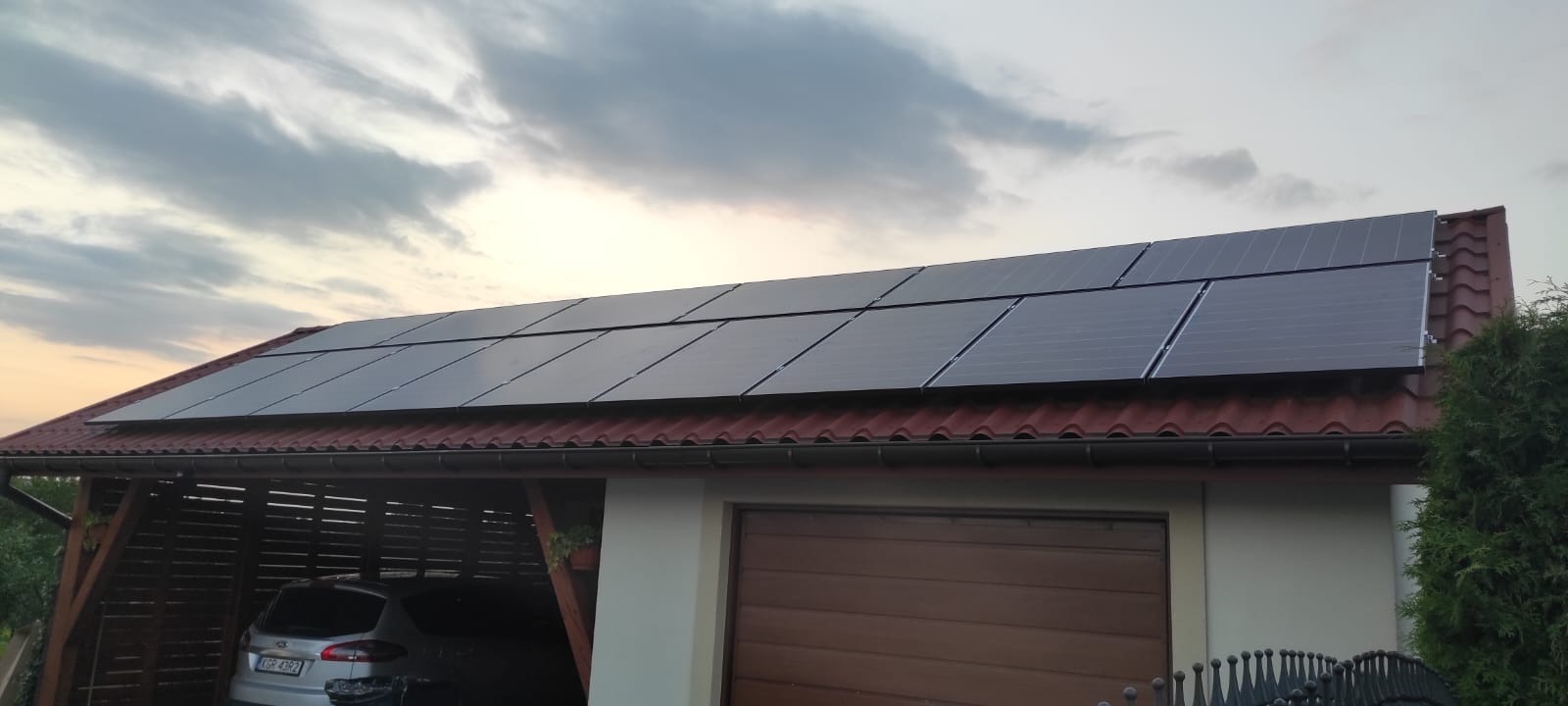 Panele słoneczne zamontowane na dachu garażu z czerwoną dachówką, widoczne auto zaparkowane pod wiatą, pochmurne niebo w tle.
