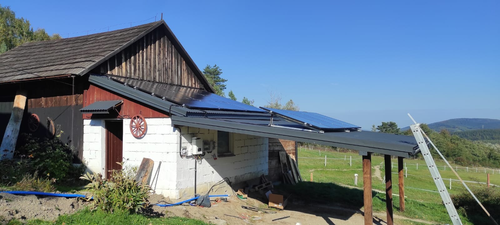 Instalacja paneli słonecznych na dachu budynku gospodarczego z drewnianymi elementami, widoczne okablowanie i skrzynka elektryczna na ścianie, w tle zalesione wzgórza.