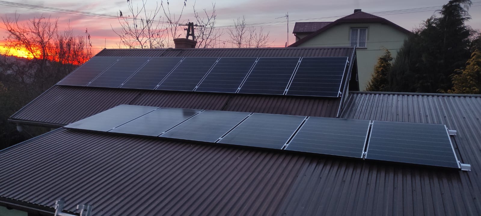 Panele fotowoltaiczne zamontowane na brązowym, blaszanym dachu domu jednorodzinnego, widoczne na tle zachodzącego słońca i drzew bez liści.