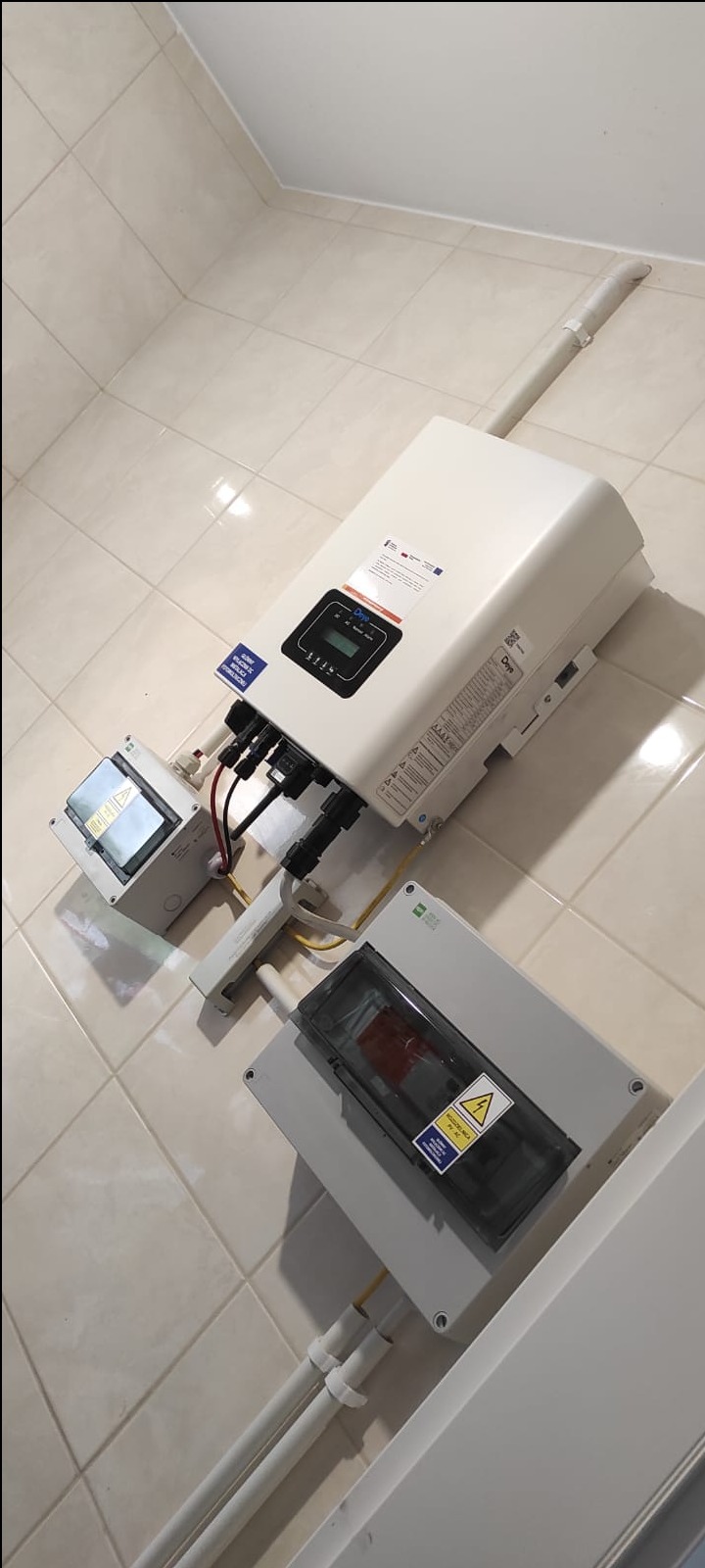 Biały inwerter fotowoltaiczny zamontowany na jasnej, kafelkowanej ścianie, z widocznymi kablami i skrzynkami przyłączeniowymi.