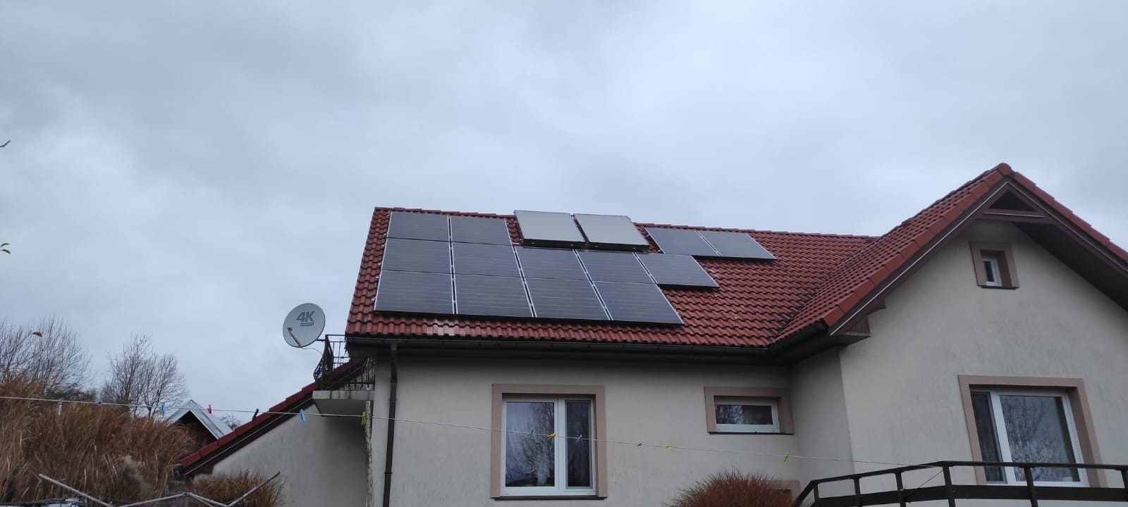 Dom jednorodzinny z panelami słonecznymi na dachu z czerwonej dachówki, widoczne okna i antena satelitarna, pochmurne niebo.