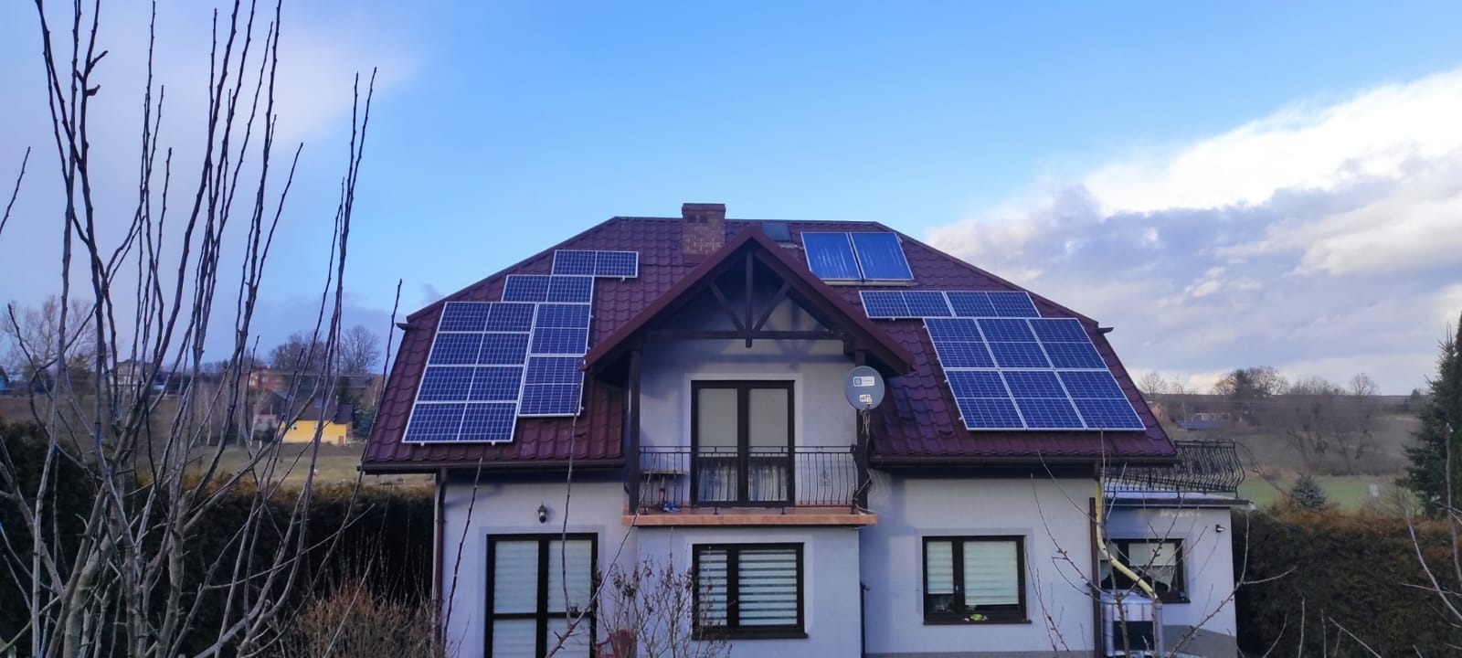 Dom jednorodzinny z panelami słonecznymi na dachu pokrytym czerwoną dachówką, widok zza drzew bez liści w pochmurny dzień.