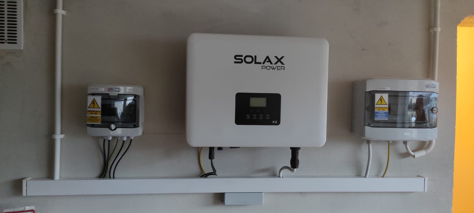 Biały inwerter Solax Power X3 zamontowany na ścianie obok dwóch skrzynek z zabezpieczeniami, widoczne kable i przewody.