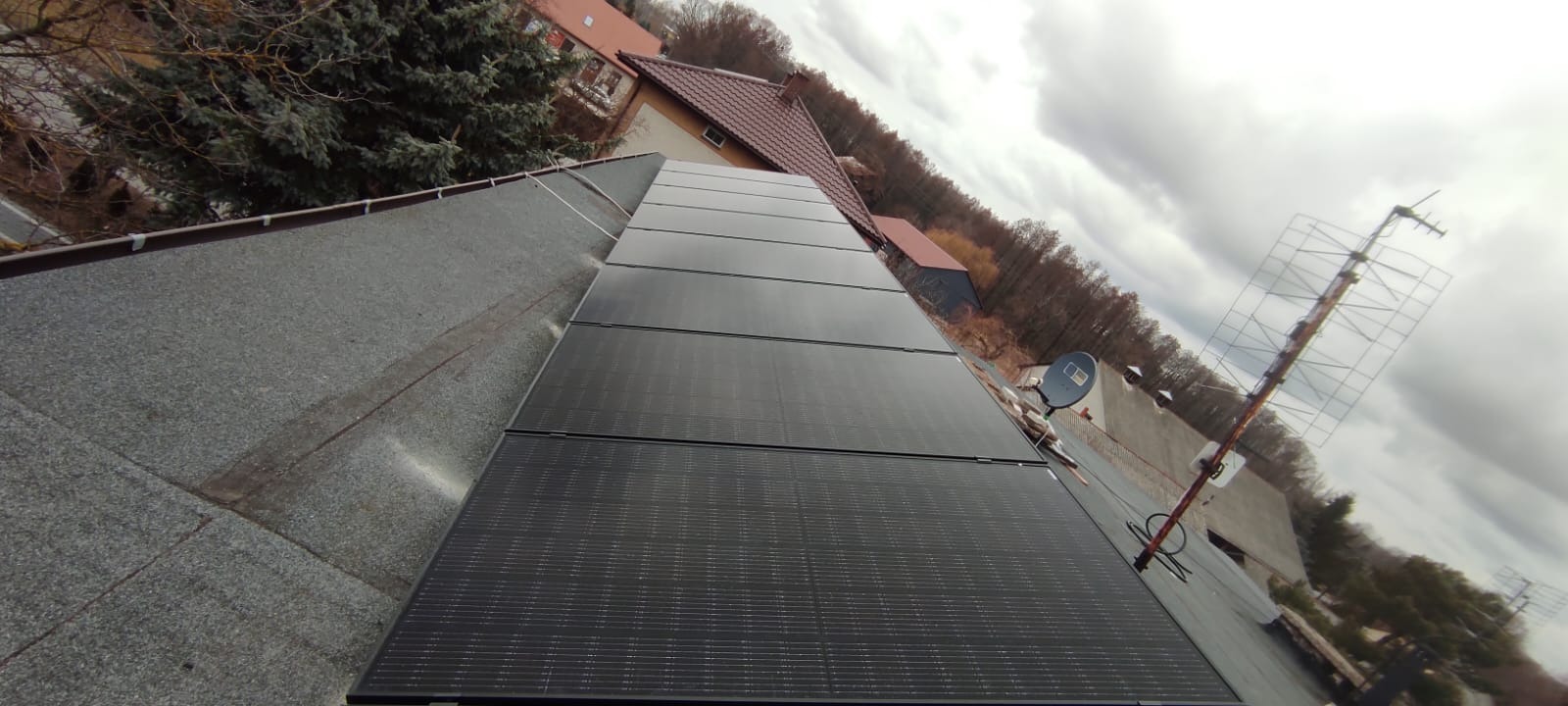Panele fotowoltaiczne na dachu pokrytym papą, widoczne budynki mieszkalne w tle oraz antena telewizyjna.
