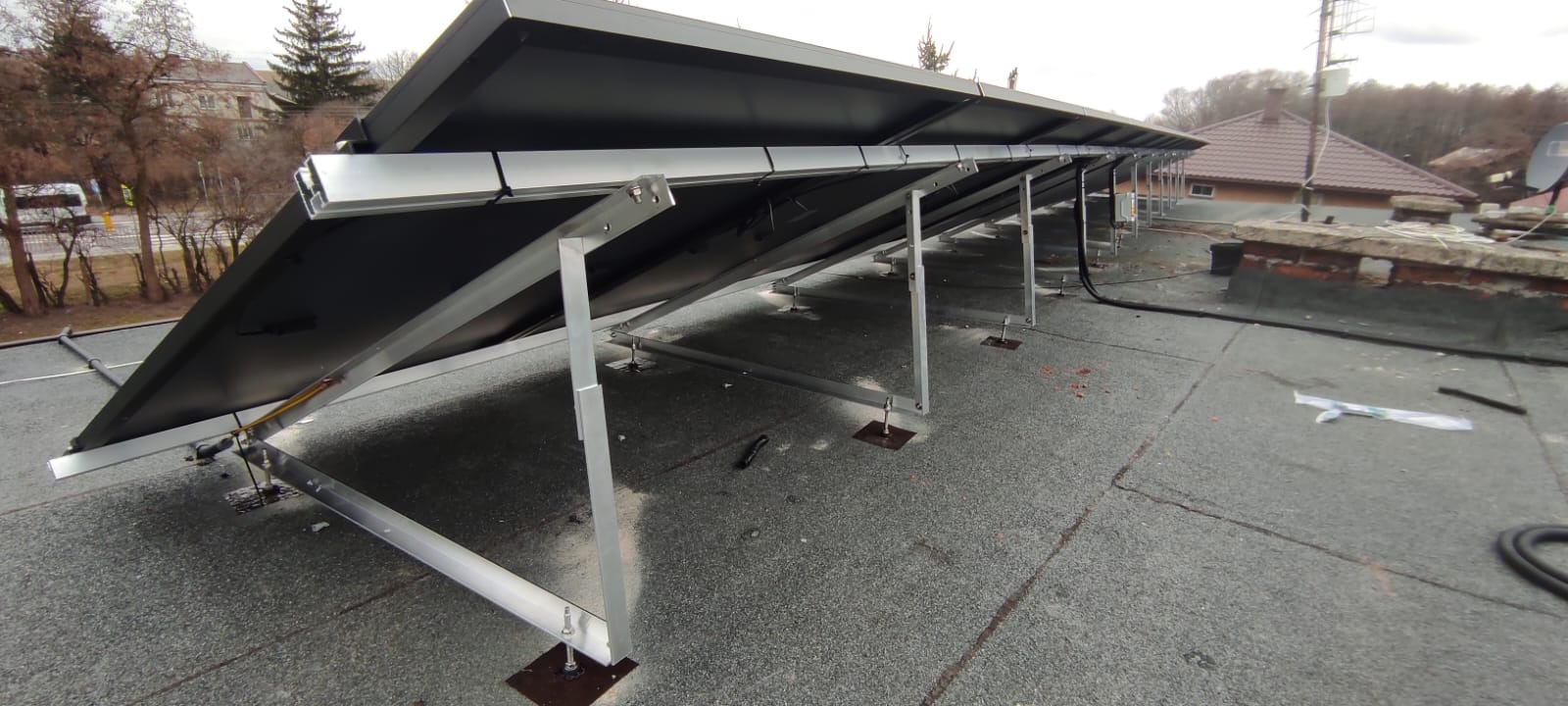 Panele słoneczne na płaskim dachu z widocznymi elementami montażowymi, w tle budynki mieszkalne i drzewa.