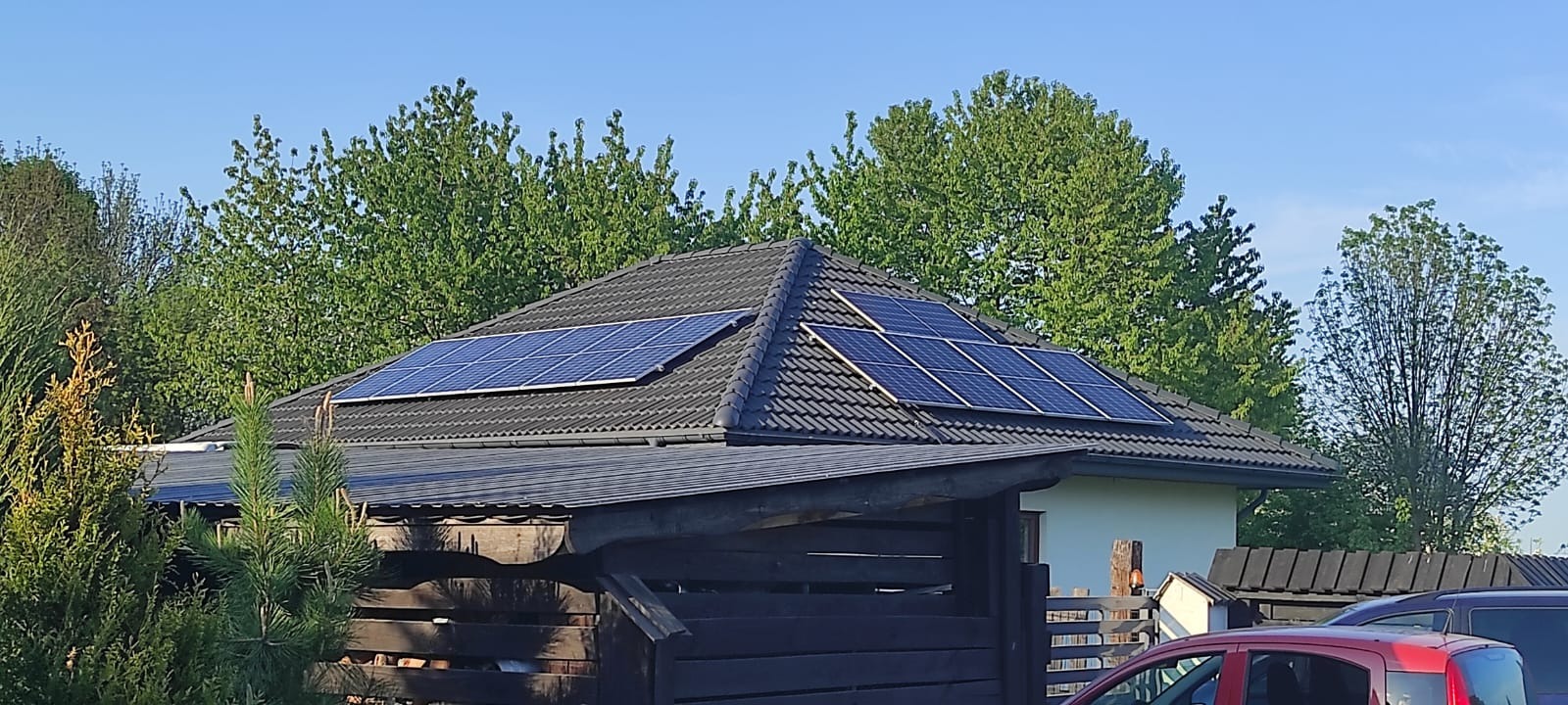 Instalacja paneli słonecznych na dachu domu jednorodzinnego z ciemnej dachówki, widok z poziomu gruntu przez drewniane ogrodzenie.