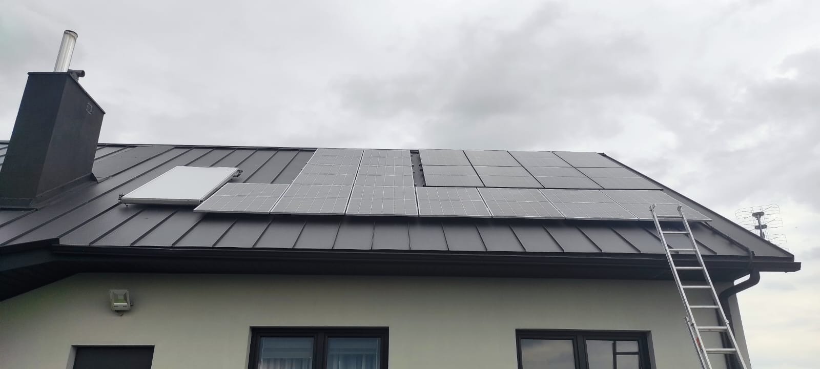 Montaż paneli słonecznych na dachu z blachy trapezowej, widoczna drabina oparta o dach oraz antena telewizyjna w tle.