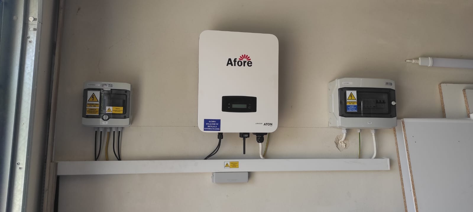 Biały inwerter fotowoltaiczny marki Afore zamontowany na ścianie obok dwóch szarych skrzynek elektrycznych z żółtymi tabliczkami ostrzegawczymi, połączonych białą listwą.
