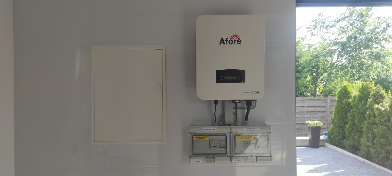 Biały inwerter Afore zamontowany na kafelkowanej ścianie, z dwoma licznikami energii poniżej, obok skrzynki elektrycznej i z widokiem na ogród przez otwarte drzwi.