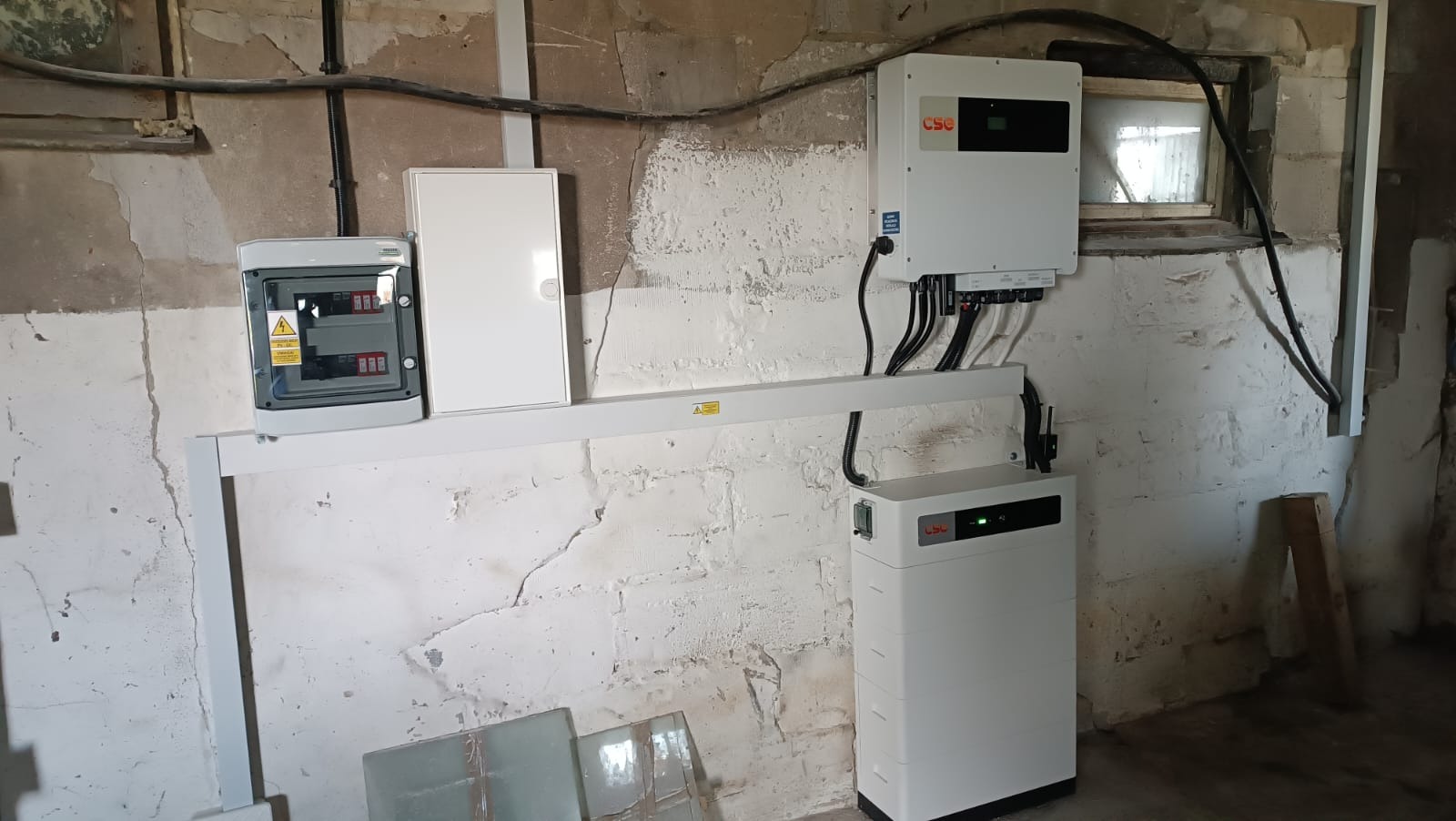 Instalacja wewnętrzna z akumulatorem, falownikiem i rozdzielnicą na tle surowej ściany z widocznymi instalacjami elektrycznymi.