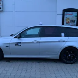 Srebrne kombi taxi z czarnymi felgami i niebieskimi zaciskami hamulcowymi, postój przy lokalu gastronomicznym z reklamami sezonowymi.