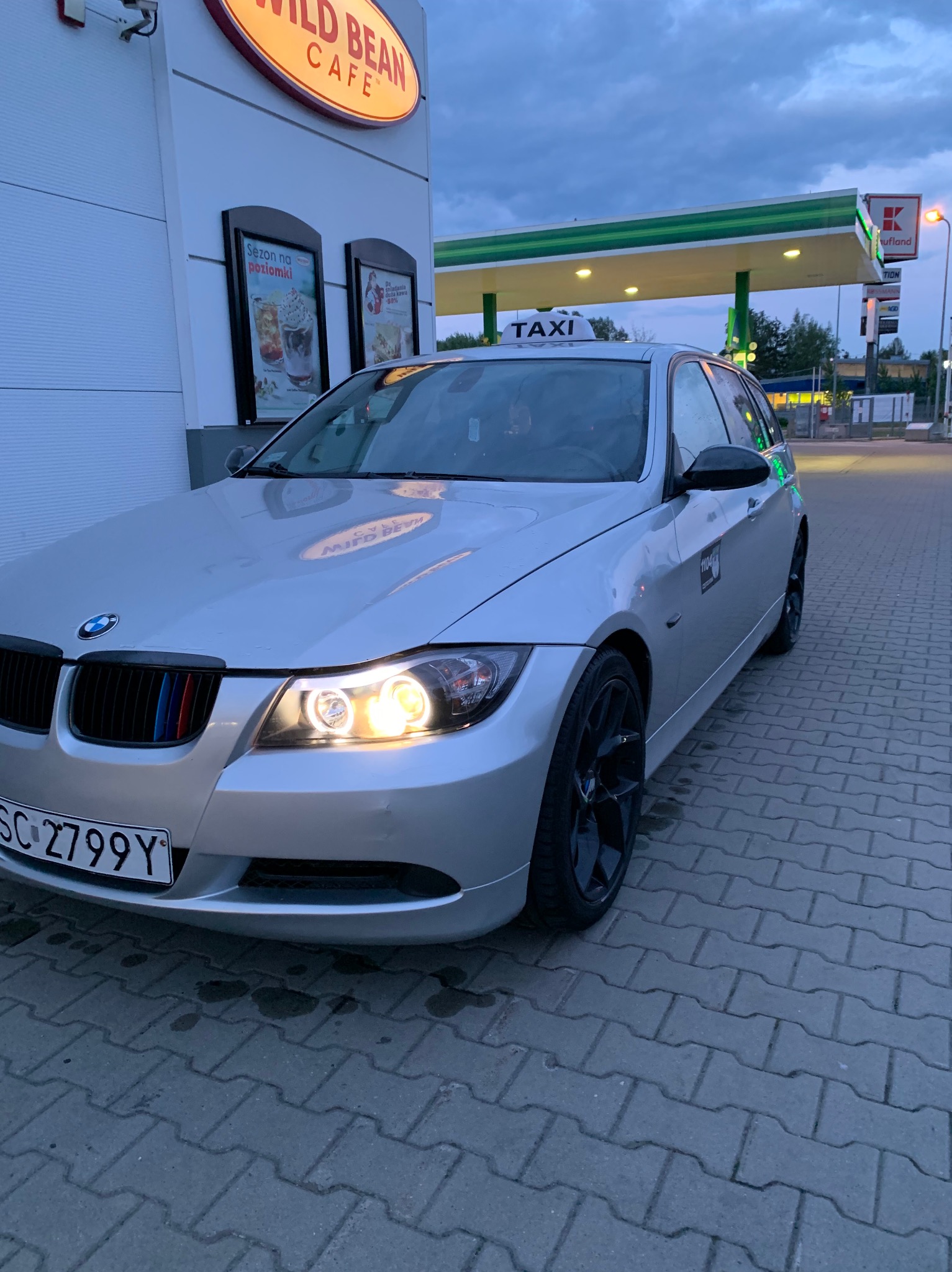 Srebrne BMW kombi z napisem TAXI na dachu, zaparkowane na stacji benzynowej z oświetlonymi reflektorami i czarnymi felgami.