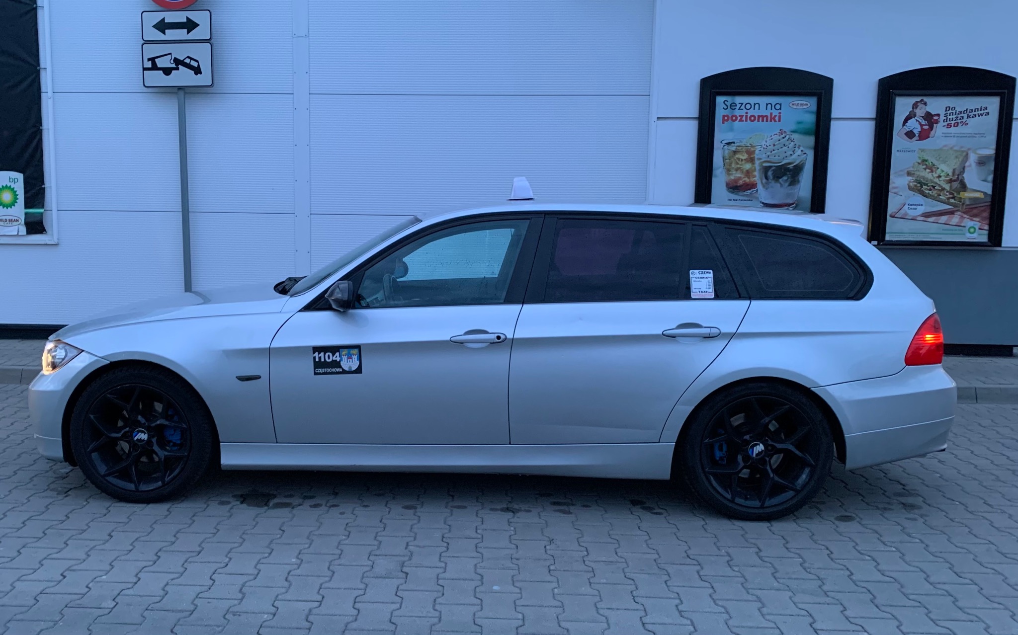 Srebrne kombi taxi z czarnymi felgami i niebieskimi zaciskami hamulcowymi, postój przy lokalu gastronomicznym z reklamami sezonowymi.