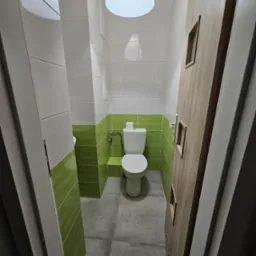 Wnętrze łazienki z toaletą, wykończone białymi i zielonymi płytkami, szarą podłogą oraz drewnianymi drzwiami z minimalistycznymi przeszkleniami.