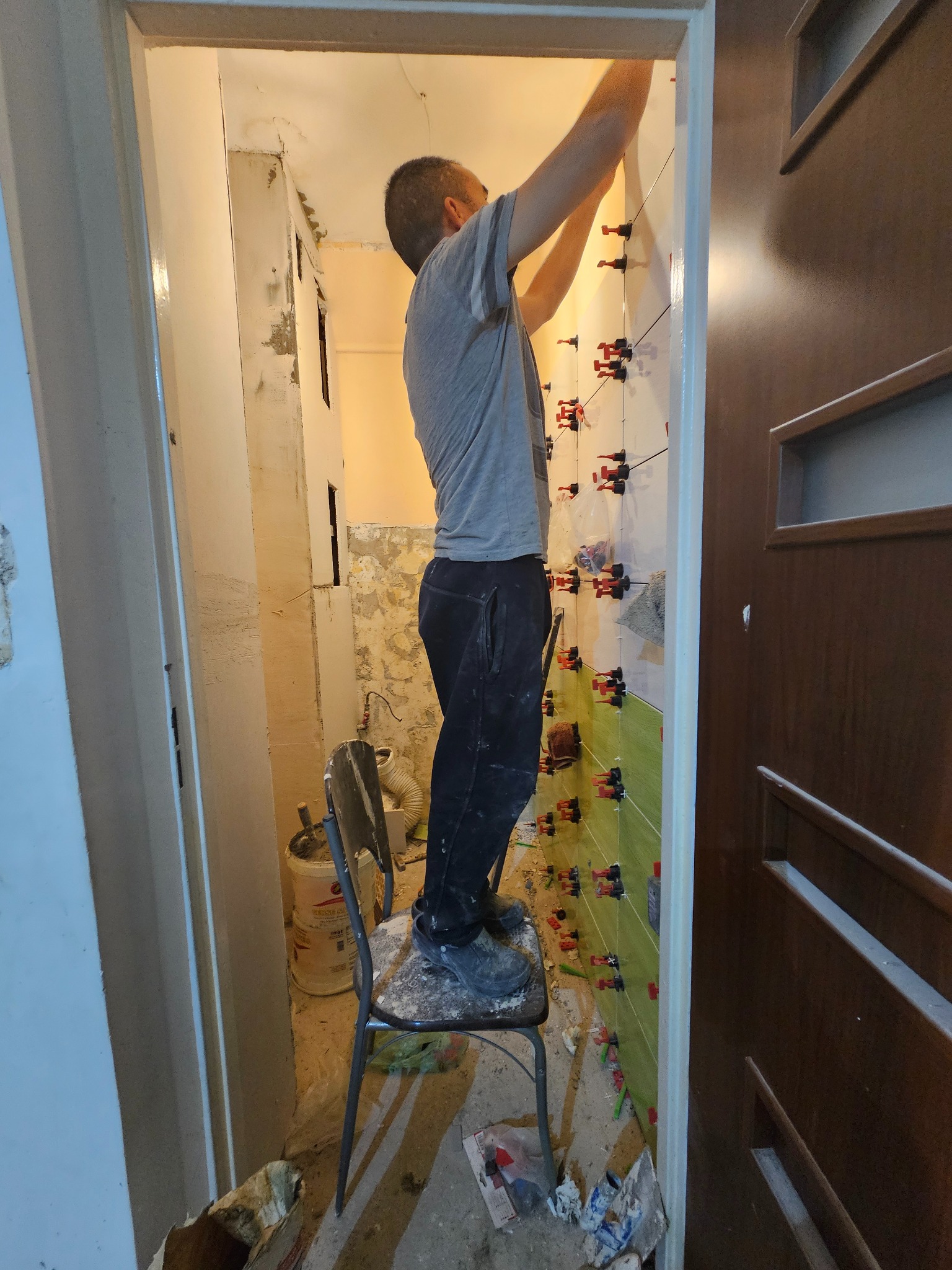 Mężczyzna na krześle układa zielone płytki w łazience, używając systemu poziomowania płytek, widoczne ślady wcześniejszych prac na ścianach.