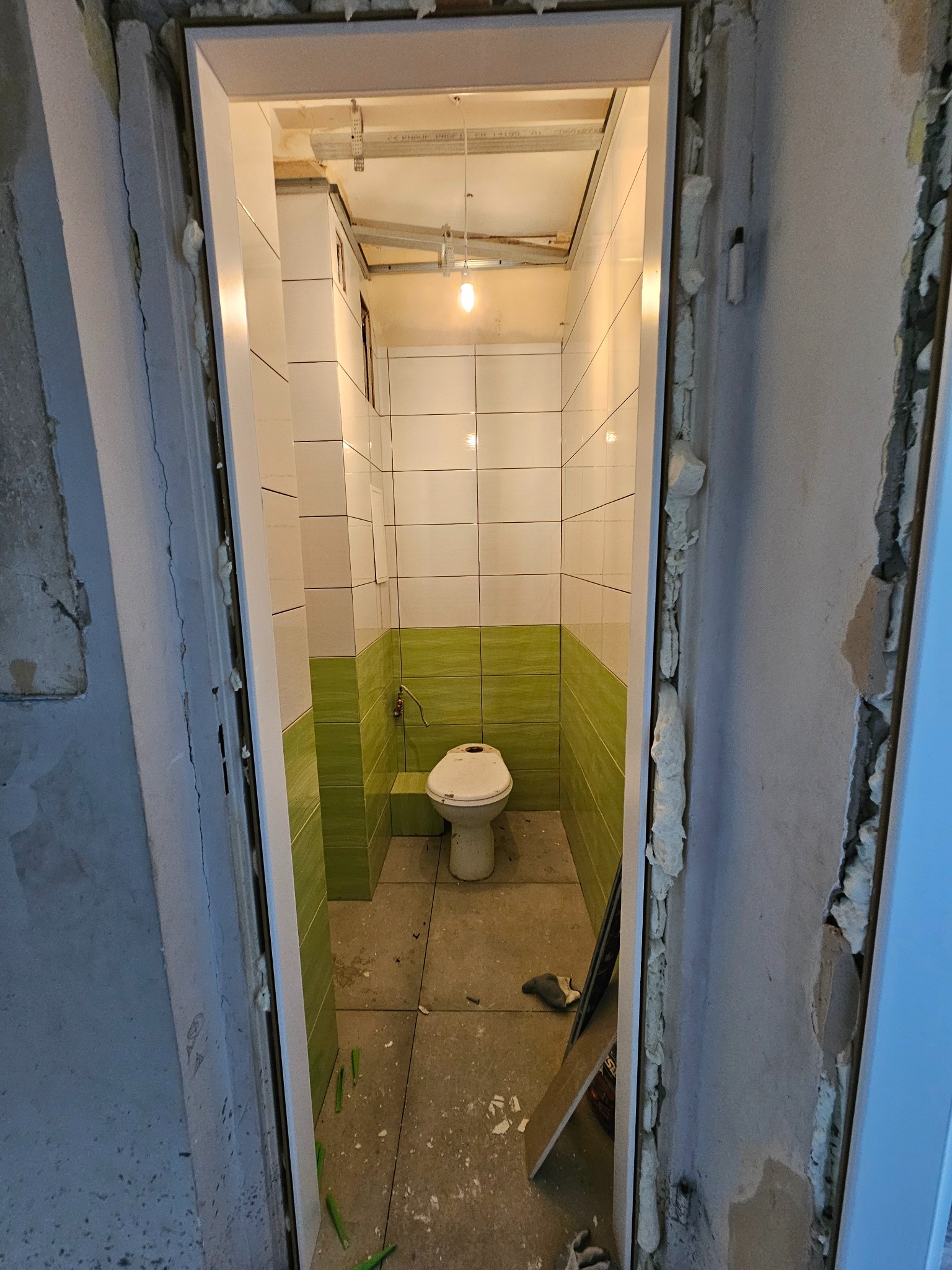 Wnętrze remontowanej łazienki z biało-zielonymi płytkami, widok przez niedokończone futryny drzwiowe z pianką montażową, sufit w trakcie prac wykończeniowych.